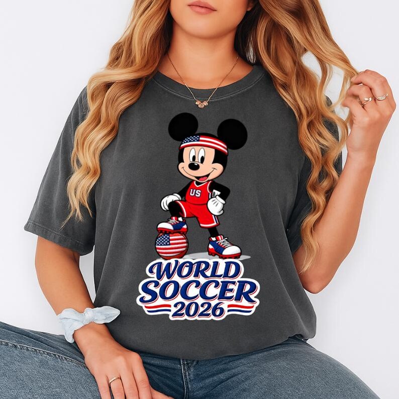 May include: Dark gray t-shirt featuring Mickey Mouse in a soccer uniform with a US flag headband. Mickey is standing on a soccer ball with a US flag design. The text "WORLD SOCCER 2026" is below the character in red, white, and blue.