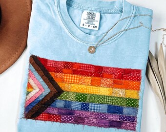 May include: A light blue t-shirt featuring a rectangular patch with a pride flag design made of various fabric squares in red, orange, yellow, green, blue, purple, brown, and pink. The shirt has a Comfort Colors label and a silver pendant necklace.
