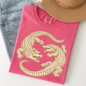May include: A pink t-shirt featuring a detailed illustration of two alligators facing each other. The alligators are in a light brown color. The shirt is folded and placed next to a pair of blue jeans and a brown hat.