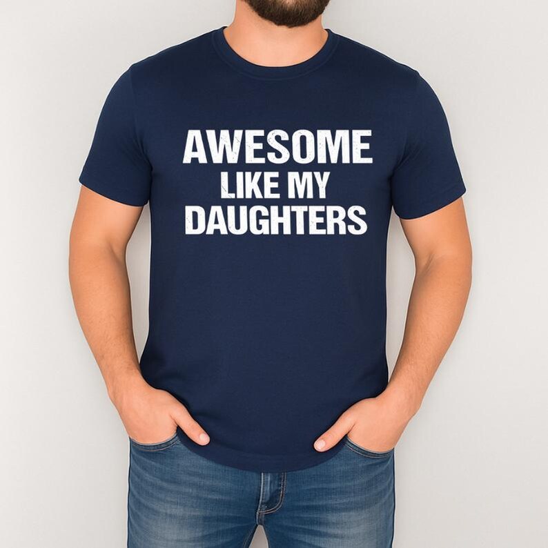 May include: Navy blue t-shirt with the white text "AWESOME LIKE MY DAUGHTERS". The shirt is made of a soft material and has a crew neck. The person in the image is wearing blue jeans.