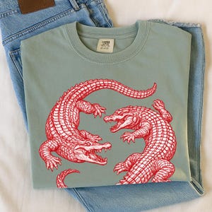 May include: Sage green t-shirt featuring a red illustration of two alligators facing each other. The t-shirt is folded and placed on top of a pair of blue jeans. The t-shirt has a crew neck and short sleeves. The image includes a small tag.
