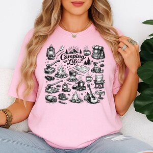 May include: A light pink t-shirt featuring a black and white camping-themed graphic. The graphic includes illustrations of camping gear such as a lantern, campfire, axe, backpack, and a guitar. The words "Camping Life" are also displayed.