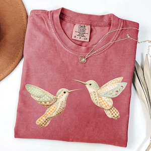 May include: A dusty rose Comfort Colors t-shirt featuring two hummingbird appliqués with patchwork wings. The shirt has a crew neck and short sleeves. A silver necklace with a small pendant is draped on the shirt.