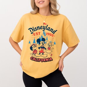 May include: Mustard yellow t-shirt with a Disneyland graphic. The design features Mickey Mouse, Minnie Mouse, and Donald Duck, with the text "Disneyland EST 1955 California". The shirt has short sleeves and a loose fit.