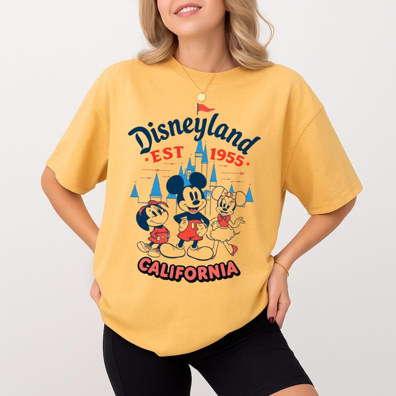 May include: Mustard yellow t-shirt with a Disneyland graphic. The design features Mickey Mouse, Minnie Mouse, and Donald Duck, with the text "Disneyland EST 1955 California". The shirt has short sleeves and a loose fit.