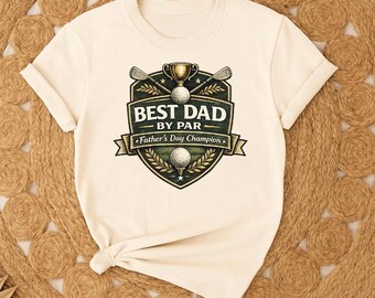 May include: A cream-colored short-sleeve t-shirt featuring a graphic of a shield emblem with two golf clubs, a trophy, and two golf balls. The text reads BEST DAD BY PAR and Father's Day Champion in a dark green and gold color scheme.