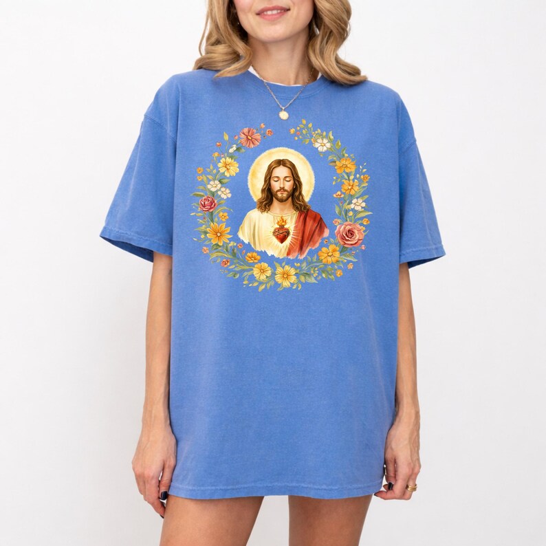 K&ouml;nnte beinhalten: Ein blaues T-Shirt mit einer Aquarellillustration von Jesus Christus, umgeben von einem Blumenkranz. Das Kunstwerk enth&auml;lt einen Heiligenschein und ein heiliges Herz. Das Shirt ist einfarbig blau und hat kurze &Auml;rmel.