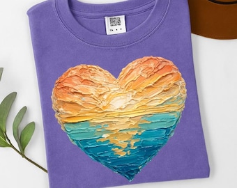 May include: A purple t-shirt featuring a heart-shaped design. The heart is divided into sections of orange, yellow, and blue, resembling a sunset or seascape. The shirt is folded and lies on a white surface.