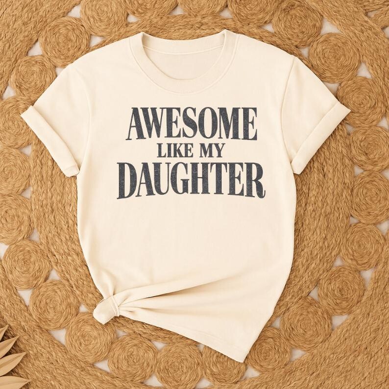 May include: A cream-coloured t-shirt with rolled sleeves. The shirt displays the text "AWESOME LIKE MY DAUGHTER" in dark grey block letters. The t-shirt is knotted at the bottom and rests on a woven, circular rug.