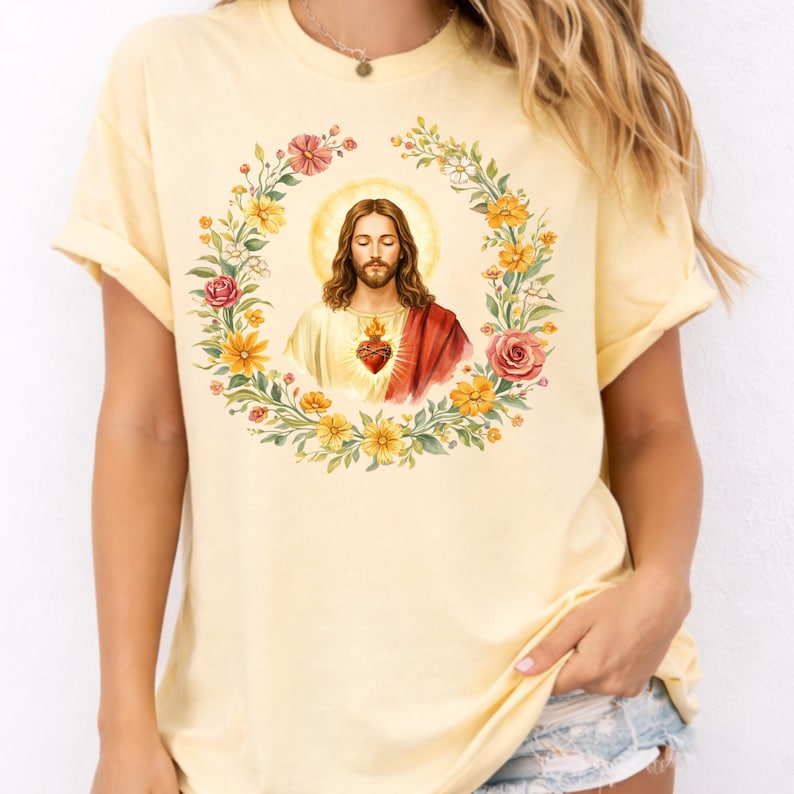 K&ouml;nnte beinhalten: Hellgelbes T-Shirt mit religi&ouml;ser Grafik. Die Grafik zeigt eine Figur mit langen braunen Haaren, einem Heiligenschein und einem roten Herzen, umgeben von einem Blumenkranz aus Gelb, Rosa und Gr&uuml;n.