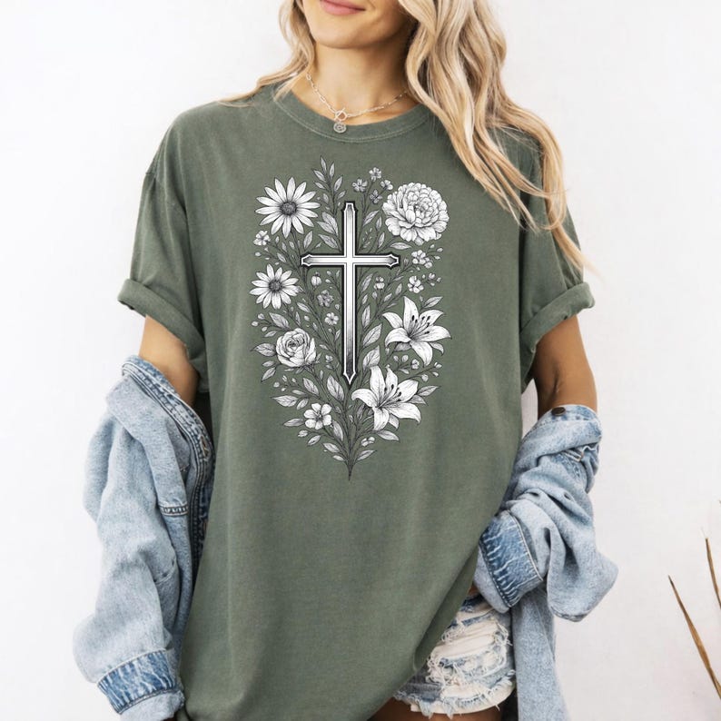 Op de afbeelding: Olijfgroen T-shirt met een centraal ontwerp van een kruis omringd door witte bloemen en bladeren. De afbeelding is gedetailleerd en het T-shirt heeft korte mouwen en een losse pasvorm. De kleur is een gedempt groen.