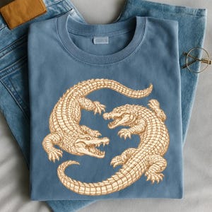 May include: A blue t-shirt featuring a detailed illustration of two alligators facing each other. The alligators are tan with textured scales and open mouths. The shirt is folded on top of blue denim jeans.