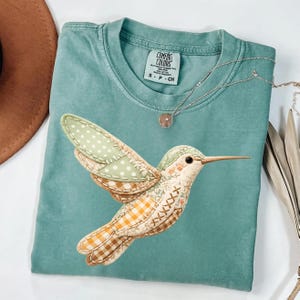 May include: A teal Comfort Colors t-shirt featuring a detailed hummingbird applique with patchwork wings and body. The hummingbird has a long beak and is stitched with various patterns. A silver necklace with a pendant is also visible.