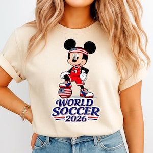 May include: Beige t-shirt featuring Mickey Mouse in a soccer uniform with a soccer ball. Mickey is wearing a red and white uniform with "US" on the chest. The text "WORLD SOCCER 2026" is below the character.