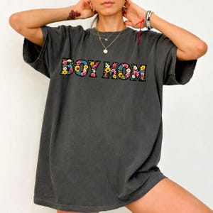 May include: Dark gray oversized t-shirt with the text "BOY MOM" in colorful floral letters. The shirt has short sleeves and a relaxed fit, perfect for casual wear. The floral design includes pink, yellow, and blue flowers.