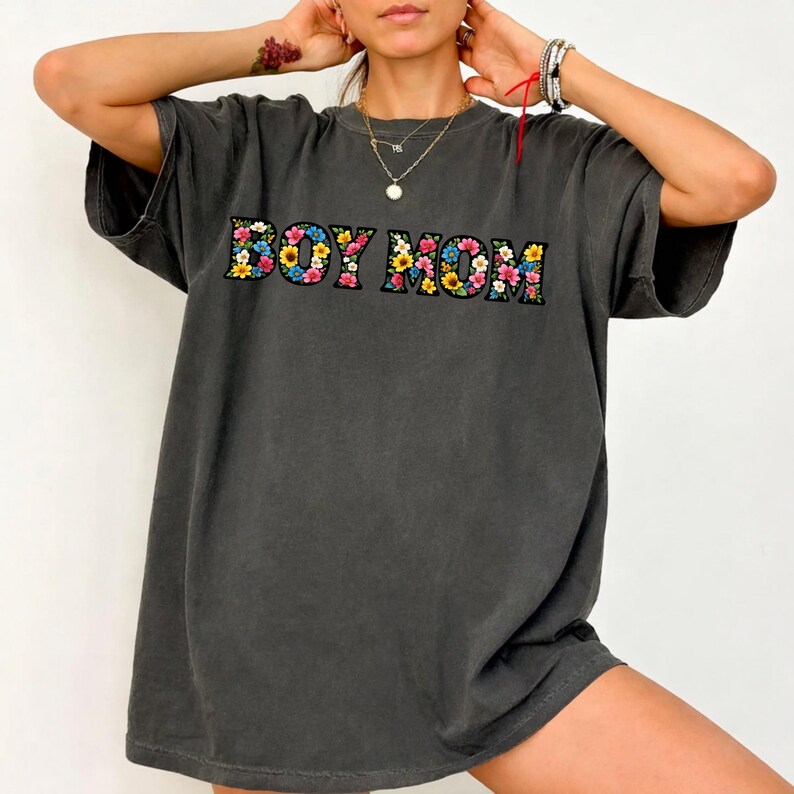 May include: Dark gray oversized t-shirt with the text "BOY MOM" in colorful floral letters. The shirt has short sleeves and a relaxed fit, perfect for casual wear. The floral design includes pink, yellow, and blue flowers.