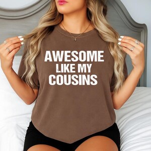 May include: A brown t-shirt with the white text "AWESOME LIKE MY COUSINS". The shirt is a short-sleeved crew neck style. The model is wearing black shorts and jewelry.