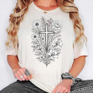 Op de afbeelding: Cr&egrave;mekleurig T-shirt met een gedetailleerd zwart-wit bloemenontwerp met een kruis in het midden. Het ontwerp bevat verschillende bloemen en bladeren, wat een botanisch en religieus thema cre&euml;ert. Het shirt heeft korte mouwen en een ronde hals.
