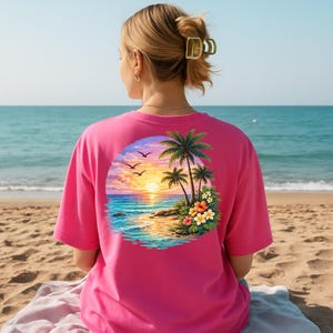 May include: A bright pink t-shirt with a tropical sunset design on the back. The graphic shows palm trees, ocean waves, and a colourful sky with birds. The shirt is short-sleeved and made of a soft material.