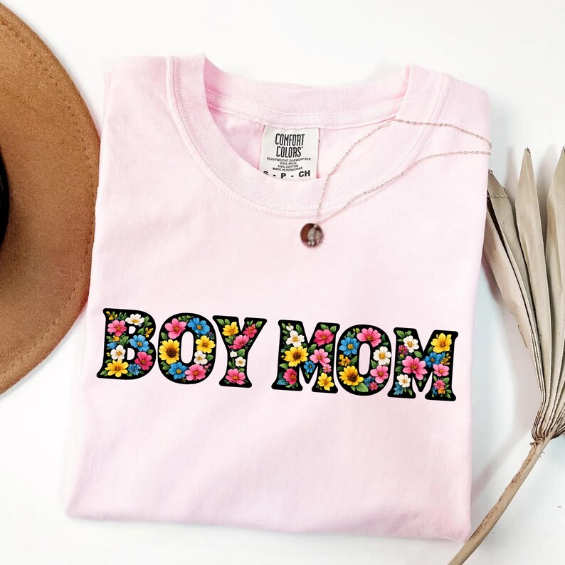 May include: A light pink t-shirt with the words "BOY MOM" in a floral design. The shirt has a crew neck and short sleeves. A gold necklace with a pendant is on the shirt.