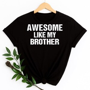 May include: Black t-shirt with the white text "AWESOME LIKE MY BROTHER." The shirt is hanging on a wooden hanger. A sprig of greenery is in the lower left corner.