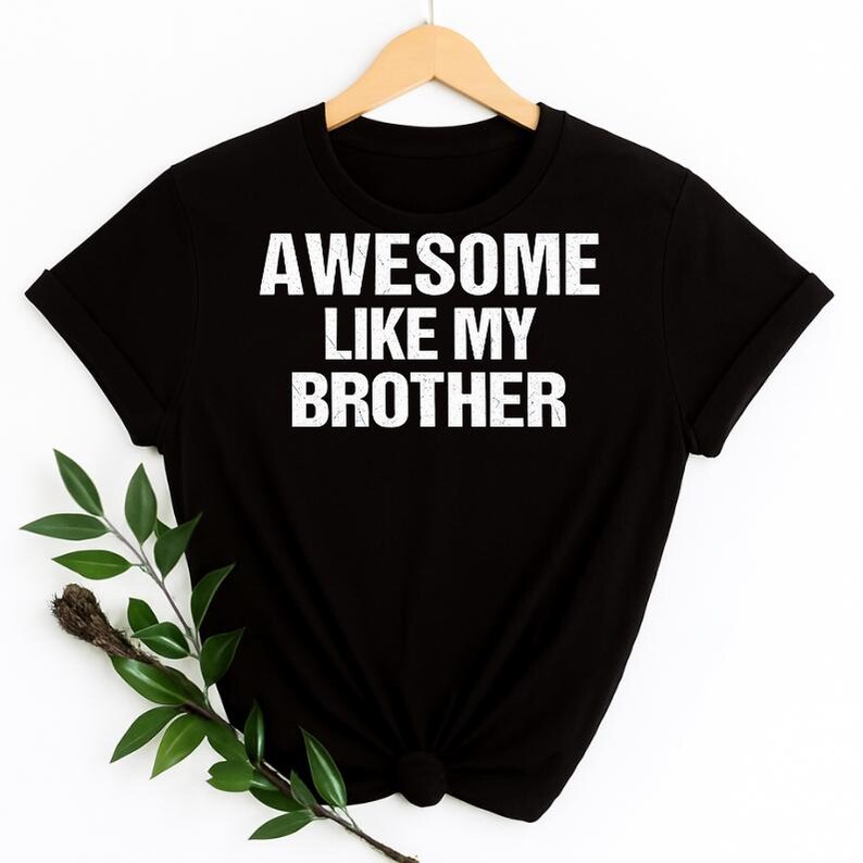 May include: Black t-shirt with the white text "AWESOME LIKE MY BROTHER." The shirt is hanging on a wooden hanger. A sprig of greenery is in the lower left corner.