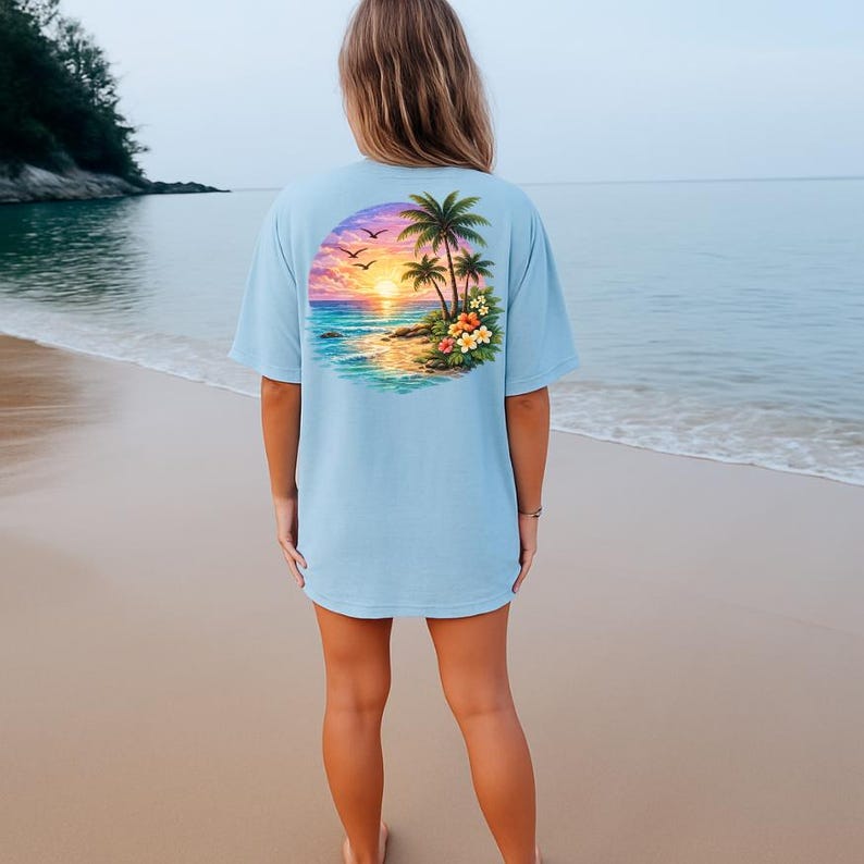 May include: Pale blue t-shirt with a tropical sunset graphic on the back. The design shows palm trees, flowers, and birds set against a colourful sky and ocean. The shirt is a relaxed, casual style, suitable for everyday wear.