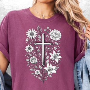 Op de afbeelding: Een bordeauxrood T-shirt met een gedetailleerde illustratie van een kruis omringd door witte bloemen en bladeren. Het ontwerp omvat madeliefjes, lelies en rozen, waardoor een bloemrijk en religieus thema ontstaat. Het kruis is gecentreerd, met de bloemen eromheen gerangschikt.