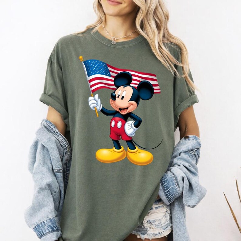 Puede incluir: Camiseta verde oliva con un dibujo de Mickey Mouse sosteniendo la bandera estadounidense. Mickey lleva pantalones cortos rojos, zapatos amarillos y guantes blancos. La bandera tiene rayas rojas y blancas con un campo de estrellas azul.