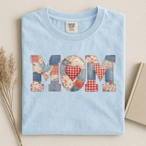 May include: A light blue Comfort Colors t-shirt with the word "MOM" appliqued on the front. The letters are made of patchwork fabric in various patterns, including denim, floral, and gingham. A red heart is in the center of the "O".