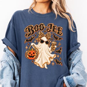 May include: A slate blue t-shirt featuring a graphic of a ghost wearing leopard print sunglasses and a matching bow. The ghost holds a leopard print cup and a jack-o'-lantern pumpkin. The text BOO-JEE is printed above, surrounded by small bats and stars.
