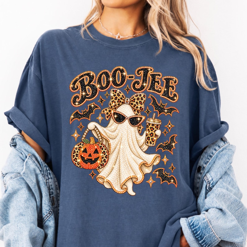 May include: A slate blue t-shirt featuring a graphic of a ghost wearing leopard print sunglasses and a matching bow. The ghost holds a leopard print cup and a jack-o'-lantern pumpkin. The text BOO-JEE is printed above, surrounded by small bats and stars.