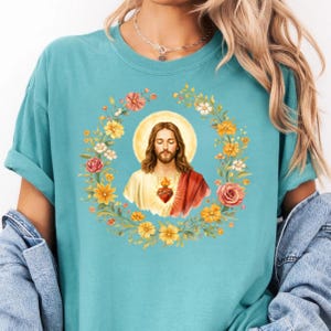 K&ouml;nnte beinhalten: Ein hellblaues T-Shirt mit einem religi&ouml;sen Bild von Jesus, umgeben von einem Blumenkranz. Das Bild zeigt Jesus mit Heiligenschein, gekleidet in ein wei&szlig;es Gewand und einen roten Schal, mit einem Herzen auf der Brust. Der Kranz enth&auml;lt gelbe, rosa und wei&szlig;e Blumen.