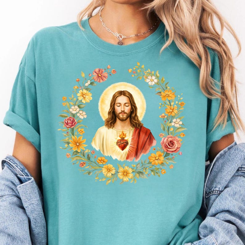 K&ouml;nnte beinhalten: Ein hellblaues T-Shirt mit einem religi&ouml;sen Bild von Jesus, umgeben von einem Blumenkranz. Das Bild zeigt Jesus mit Heiligenschein, gekleidet in ein wei&szlig;es Gewand und einen roten Schal, mit einem Herzen auf der Brust. Der Kranz enth&auml;lt gelbe, rosa und wei&szlig;e Blumen.