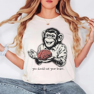 May include: Cream-colored t-shirt featuring a smiling chimpanzee holding a red brain. The chimpanzee is wearing a white shirt. The text "you should use your brain!" is printed below the graphic.