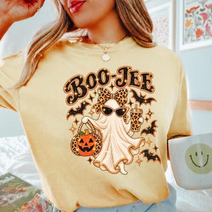 May include: A pale yellow t-shirt featuring a graphic of a ghost wearing a leopard print bow and sunglasses, holding a leopard print pumpkin bucket and a matching cup. The text BOO-JEE is printed above the ghost, surrounded by black bats.
