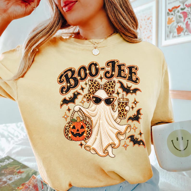 May include: A pale yellow t-shirt featuring a graphic of a ghost wearing a leopard print bow and sunglasses, holding a leopard print pumpkin bucket and a matching cup. The text BOO-JEE is printed above the ghost, surrounded by black bats.