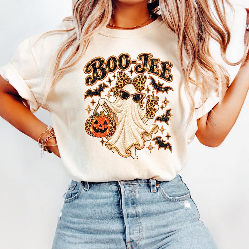 May include: A cream-colored t-shirt featuring a graphic of a ghost wearing sunglasses and a leopard print bow, holding a pumpkin and a drink. The text BOO-JEE is printed above the ghost, surrounded by leopard print bats and star accents.