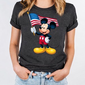 Puede incluir: Camiseta gris oscuro con Mickey Mouse sosteniendo una bandera estadounidense. Mickey lleva pantalones cortos rojos, zapatos amarillos y guantes blancos. La bandera tiene rayas rojas y blancas y un campo azul con estrellas blancas.