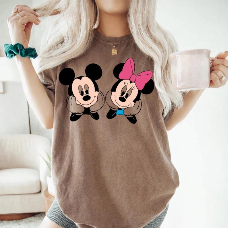 May include: Brown t-shirt featuring a graphic of Mickey and Minnie Mouse. Mickey is wearing a black bow tie and Minnie is wearing a pink bow. The cartoon characters are smiling and resting their chins on their hands.