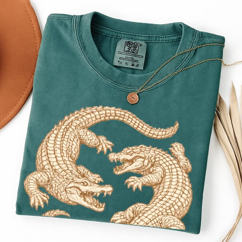 May include: Teal-colored t-shirt featuring a detailed illustration of two alligators in a fighting pose. The alligators are in a light brown color. A gold necklace with a circular pendant is draped over the shirt.
