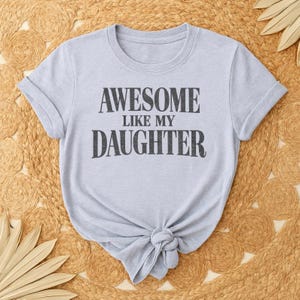 May include: A pale blue t-shirt featuring the text "AWESOME LIKE MY DAUGHTER" in dark grey lettering. The shirt is knotted at the hem and has rolled sleeves. It is presented on a woven, tan surface.