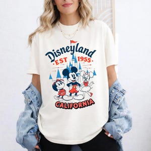 May include: Cream-coloured t-shirt with a Disneyland graphic. The design features Mickey Mouse, Minnie Mouse, and Donald Duck, with the text "Disneyland EST 1955 CALIFORNIA" in red and blue. The shirt has short sleeves and a relaxed fit.