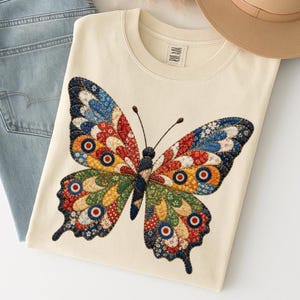 May include: Cream-colored sweatshirt featuring a large, colorful butterfly design. The butterfly's wings are composed of various patterned fabrics in shades of red, blue, yellow, and green. The design includes circular accents. A pair of blue jeans is visible.