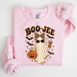 Pu&ograve; includere: Una felpa girocollo rosa chiaro con la grafica di un fantasma che indossa un fiocco e occhiali da sole leopardati. Il fantasma tiene in mano un bicchiere leopardato e un secchiello a forma di zucca. Il testo BOO-JEE &egrave; stampato sopra con pipistrelli.