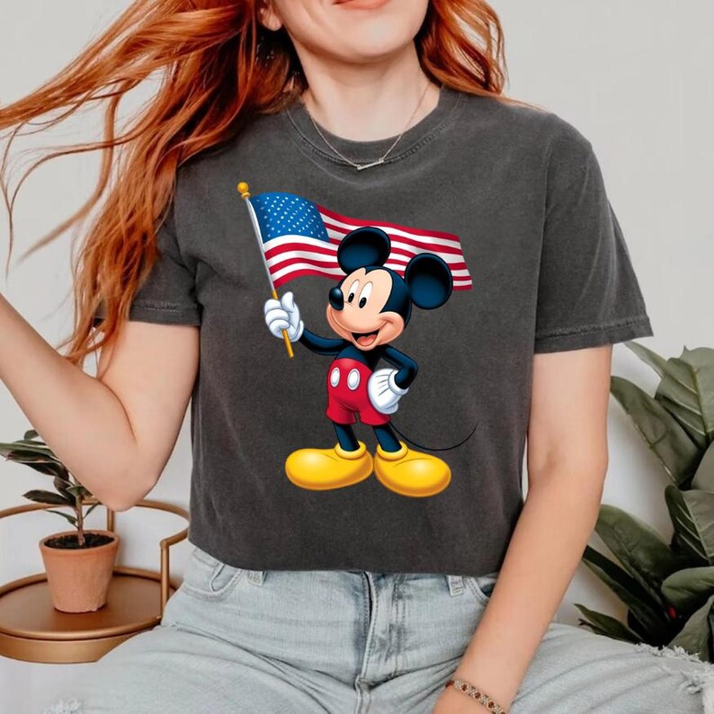 Puede incluir: Camiseta gris oscuro con un gr&aacute;fico de Mickey Mouse sosteniendo una bandera estadounidense. Mickey lleva pantalones cortos rojos, zapatos amarillos y guantes blancos. La bandera estadounidense tiene rayas rojas y blancas y un campo azul de estrellas.