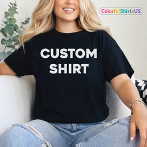 Könnte beinhalten: Schwarzes T-Shirt mit den Worten "CUSTOM SHIRT" in großen, weißen Blockbuchstaben. Das T-Shirt wird von einer Person getragen. Der Text "ColorfulShirtsUS" befindet sich in einem kleinen, abgerundeten Rechteck oben rechts.