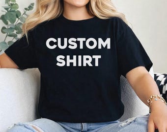 May include: Black t-shirt with the words "CUSTOM SHIRT" in large, white, block letters. The shirt is being worn by a person. The text "ColorfulShirtsUS" is in a small, rounded rectangle in the upper right corner.