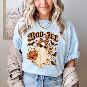 May include: A light blue t-shirt featuring a graphic of a ghost wearing a leopard print bow and sunglasses. The ghost holds a jack-o'-lantern and a drink cup, surrounded by bats and stars. The text BOO-JEE is printed in black and orange above.