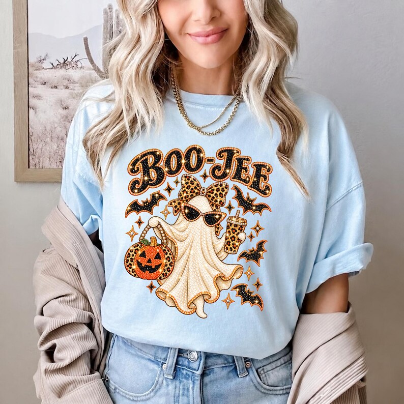 May include: A light blue t-shirt featuring a graphic of a ghost wearing a leopard print bow and sunglasses. The ghost holds a jack-o'-lantern and a drink cup, surrounded by bats and stars. The text BOO-JEE is printed in black and orange above.